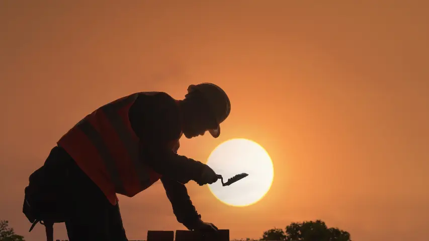 Silhouette of construction worker in hard hat and safety vest laying bricks against dramatic sunset representing temporary disability from workplace injuries in California